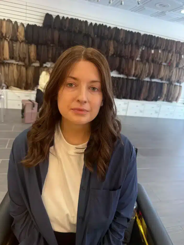 Woman with center-parted brown waves seated in front of rows of hair samples in a salon.