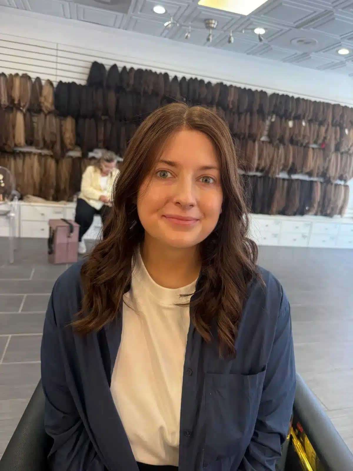 Smiling woman with long brown waves and a navy shirt seated beneath salon lights, with hair samples behind her.